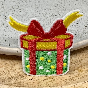May include: Embroidered patch of a green gift box with white polka dots, red ribbon, and yellow accents. The patch has a white border and is set against a neutral background.