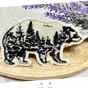 May include: A white embroidered patch featuring a black silhouette of a bear with a forest inside. The bear is standing on a wooden surface.