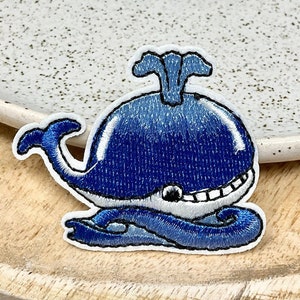 May include: A blue whale embroidered patch with a white outline. The whale is smiling and has a spout on top of its head. The whale is swimming in a blue wave.