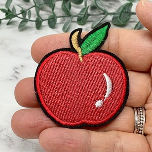 Pode incluir: Patch de maçã bordado de cor vermelha com um caule e uma folha verdes. A maçã tem um desenho bordado branco no lado.