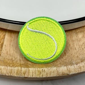 Peut inclure: Un écusson rond brodé représentant une balle de tennis. L'écusson est principalement vert néon avec une bande blanche incurvée. Il a une bordure verte et est posé sur une surface en bois, avec une assiette blanche en arrière-plan.