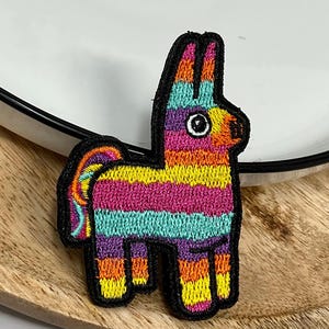 Peut inclure: Écusson brodé représentant une piñata colorée. La piñata est ornée de rayures horizontales roses, jaunes, turquoise, violettes et oranges, soulignées de noir. L'écusson est posé sur une surface en bois.