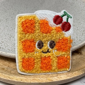 Peut inclure: Un patch brodé en forme de gaufre jaune et orange avec un visage de dessin animé mignon et deux cerises rouges sur le dessus. Le patch est sur une surface en bois.