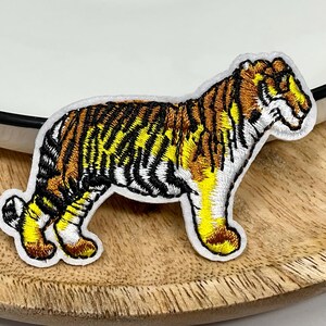 Peut inclure: Écusson brodé d'un tigre avec une bordure blanche. Le tigre présente des rayures noires, une fourrure jaune et marron, et se trouve sur une surface en bois. L'écusson est près d'une assiette blanche avec un bord noir.