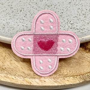Peut inclure: Patch en feutre rose en forme de pansement avec un contour blanc et un cœur rouge au centre.