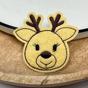 Peut inclure: Une tête de renne en feutre jaune avec des bois, des yeux et un nez marron. Le renne est brodé avec du fil noir et est placé sur un plateau en bois à côté d'une assiette blanche avec un bord noir. Le renne est une décoration festive.