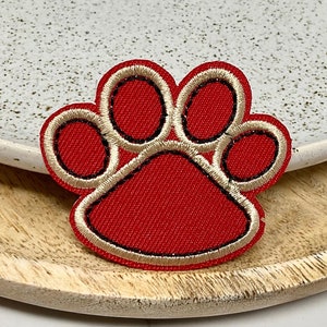 Peut inclure: Patch brodé en forme de patte de chien rouge avec un contour beige. Le patch est sur une surface en bois.