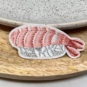 May include: A pink and white embroidered sushi patch with a shrimp design. The patch is on a wooden surface.