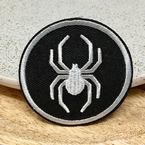 Peut inclure: Un écusson thermocollant brodé noir et blanc avec un motif d'araignée. L'araignée est blanche et le fond est noir. L'écusson est entouré d'une bordure argentée.