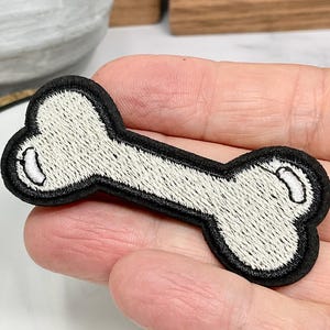 May include: A white embroidered bone-shaped patch with black stitching. The patch is on a person's hand.