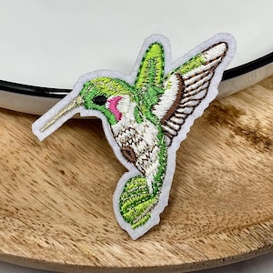 May include: An embroidered patch featuring a hummingbird in flight. The bird has vibrant green feathers on its head and back, a bright pink throat patch, white underparts, and brown-tipped wings. The patch has a white border and a pointed beak.