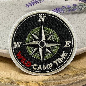 Può includere: Toppa ricamata bianca con un design a bussola nero e il testo "WILD CAMP TIME" in rosso.