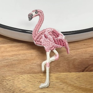 May include: An embroidered iron-on patch shaped like a standing flamingo. The bird features shades of light and dark pink thread for the body and wings, with a black and white beak and long, thin white legs standing on one foot.