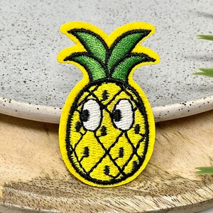 Peut inclure: Un ananas jaune avec un dessus vert feuillu et des yeux blancs. L'ananas est brodé sur un patch en tissu.