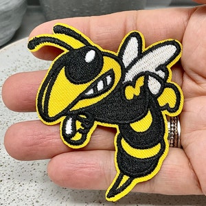 Pode incluir: Um remendo de ferro bordado de uma abelha de desenho animado com uma expressão ameaçadora, em amarelo e preto.