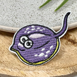 Peut inclure: Patch brodé en forme de raie, de couleur violette, avec des points blancs et de grands yeux. La raie a un contour jaune.