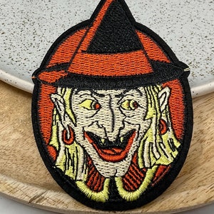 Peut inclure: Patch brodée à repasser représentant une sorcière de dessin animé avec un chapeau pointu noir, une robe orange et rouge et un sourire malicieux.
