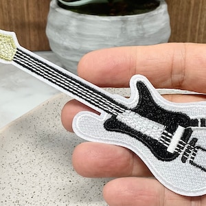 Könnte beinhalten: Weißer gestickter Aufnäher einer schwarzen E-Gitarre mit goldenem Kopf.