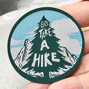 May include: A round, green embroidered patch with a white mountain scene and a pine tree in the centre. The text "GO TAKE A HIKE" is written on the tree.