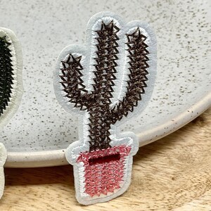Peut inclure: Deux patchs brodés de cactus, l'un vert et l'autre brun, avec des pots roses. Les patchs sont sur une surface en bois.