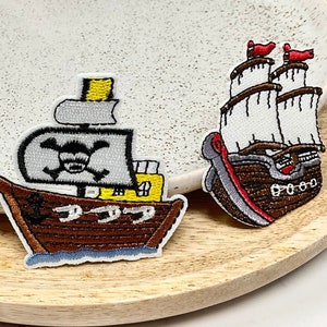 Peut inclure: Trois patchs brodés thermocollants représentant différents navires de dessin animé. Le premier navire est un voilier brun et blanc avec une voile rouge. Le deuxième navire est un navire pirate brun et jaune avec un crâne et des os croisés sur la voile. Le troisième navire est un navire à voile brun et blanc avec des accents rouges.