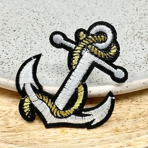 May include: White embroidered anchor with a gold rope detail. The anchor is on a white plate with a speckled surface.