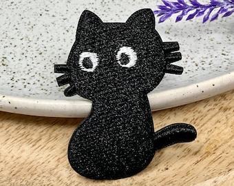 May include: A black cat patch with white eyes and whiskers. The embroidered patch is sitting on a wooden tray next to a white speckled plate and purple flowers. The cat is a cute, cartoon-like design.