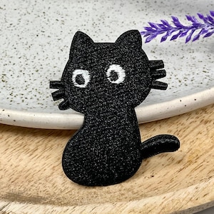Peut inclure: Un écusson de chat noir avec des yeux et des moustaches blancs. L'écusson brodé est posé sur un plateau en bois à côté d'une assiette blanche mouchetée et de fleurs violettes. Le chat a un design mignon de type dessin animé.
