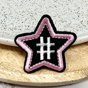 May include: Embroidered black star patch with a pink outline and a white hashtag symbol. The patch has a soft, velvety texture. The patch is placed on a wooden tray.