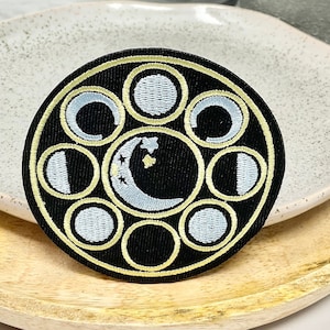 May include: A black and gold embroidered patch featuring a moon phase design. The patch depicts eight different moon phases, including a full moon, a crescent moon, and a new moon. The moon phases are encircled by a gold circle with a black background.