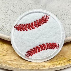 Peut inclure: Un écusson de baseball blanc et circulaire avec des détails de couture rouges. L'écusson est brodé avec le design classique des coutures de baseball. L'écusson est placé sur un plateau en bois et une assiette en céramique mouchetée.
