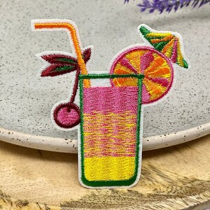 Peut inclure: Patch brodé thermocollant représentant un cocktail coloré avec une cerise, une tranche d'orange et une paille. Le cocktail est dans un grand verre avec un bord vert.