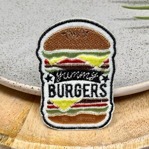 May include: A white and brown embroidered patch with a cartoon burger and the text "Yummy Burgers" in black.