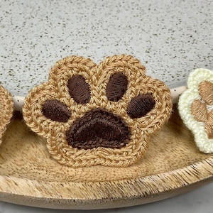 Peut inclure: Trois patchs en forme de patte de chat crochetés dans différentes couleurs. Le premier patch est marron avec une empreinte de patte blanche, le deuxième est marron avec une empreinte de patte marron et le troisième est blanc avec une empreinte de patte marron.