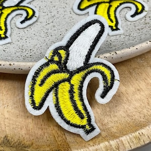 May include: Three embroidered iron-on patches of yellow bananas with black outlines. The bananas are in different positions, one is curled, one is straight, and one is partially peeled.