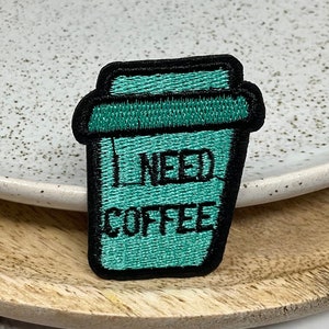 May include: An iron-on patch shaped like a coffee cup, embroidered in teal blue with the text "I NEED COFFEE" in black.