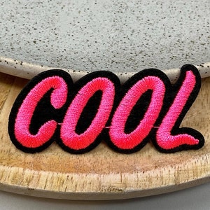 May include: Pink and black embroidered iron-on patch with the word "COOL" in a stylized font.