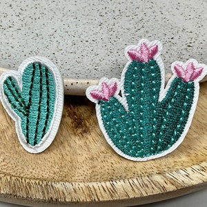 Peut inclure: Deux patchs brodés de cactus. L'un est un cactus unique avec un corps vert et une fleur rose. L'autre est un groupe de cactus avec des corps verts et des fleurs roses.