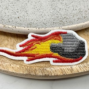 May include: Embroidered patch featuring a stylized meteor with flames. The patch has a white border, with a black and gray meteor, and yellow and red flames. The patch is on a wooden surface, with a speckled ceramic plate in the background.