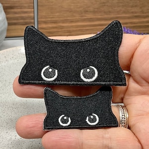 May include: Two black felt iron-on patches of a cat's face with white eyes. The larger patch is about 5 cm wide and the smaller patch is about 2.5 cm wide.
