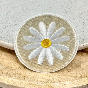 Puede incluir: Un parche de tela beige con una margarita blanca bordada. La margarita tiene un centro amarillo.