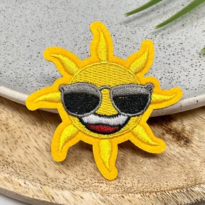 Peut inclure: Un soleil brodé jaune avec un visage souriant portant des lunettes de soleil. Le soleil est sur une surface en bois brun clair.