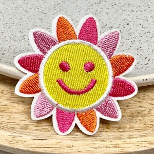 May include: A yellow smiley face sun with pink and orange flower petals. The sun is embroidered with a white outline and the flower petals are embroidered with white outlines.