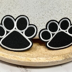 Peut inclure: Deux patchs thermocollants en forme de patte de chien noirs avec des contours blancs. Le plus grand patch est à gauche et le plus petit à droite.
