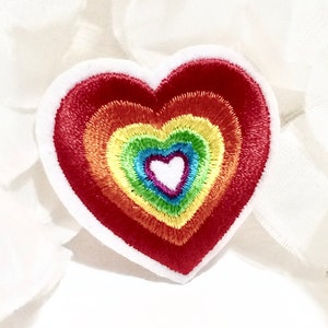 Peut inclure: Patch brodé en forme de cœur arc-en-ciel avec un cœur blanc au centre. Le patch est sur un fond blanc.