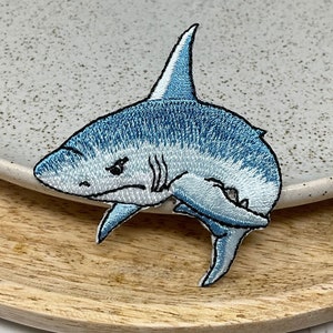 Peut inclure: Un patch brodé de requin bleu et blanc. Le requin a un ventre blanc et une nageoire dorsale bleue. Le requin est tourné vers la droite et a une expression menaçante.