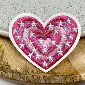 Peut inclure: Écusson brodé en forme de cœur avec une bordure blanche. Le cœur présente des couches concentriques de nuances roses et rouges, ornées d'accents blancs en forme d'étoile. Le patch est posé sur une surface en bois.