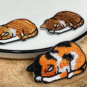 May include: Three embroidered patches of sleeping cats. One patch is orange and white, another is brown and white, and the third is calico. The patches are on a white plate and a wooden tray.