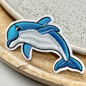 Peut inclure: Un patch brodé de dauphin bleu et blanc. Le dauphin nage vers la droite et a un ventre blanc et un dos bleu.