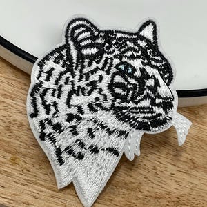 Peut inclure: Un écusson brodé représentant la tête d'un tigre blanc de profil. Le motif utilise du fil noir pour les rayures et les traits du visage du tigre, avec une touche de fil bleu pour l'œil, le tout sur un fond en tissu blanc.